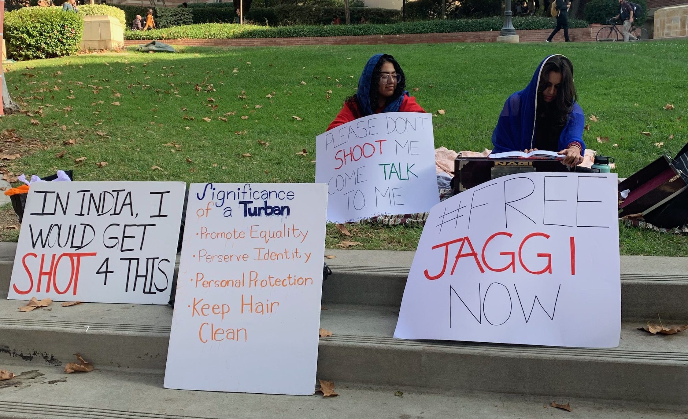 Two people sitting in grass. Posters in front of them that say: "In India, I would get shot 4 this", "significance of a turban: promote equality, perserve identity, personal protection, keep hair clean"; "please don't shoot me, come talk to me"; and "free JAGGI now"