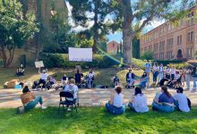 Around 23 people sit and stand around a white screen at UCLA’s Royce Quad during an accessibility protest. One person has their fist up and is holding up a sign that says “Accessibility Now! We deserve better!” Two individuals are holding a disability pride flag. Kaplan Hall and Powell Library are visible in the background.