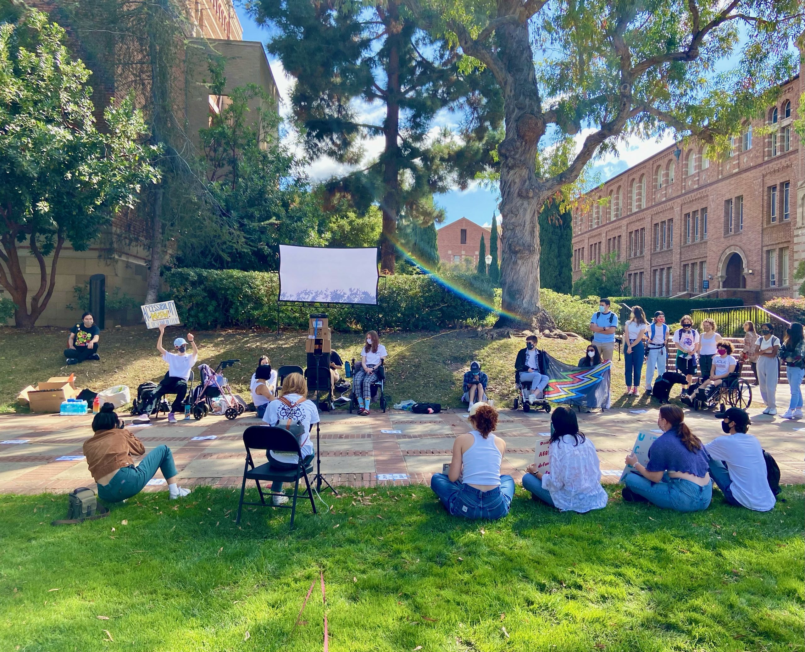 Around 23 people sit and stand around a white screen at UCLA’s Royce Quad during an accessibility protest. One person has their fist up and is holding up a sign that says “Accessibility Now! We deserve better!” Two individuals are holding a disability pride flag. Kaplan Hall and Powell Library are visible in the background.