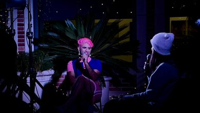 ALOK, an individual with pink hair wearing a pink and blue dress, sit in front of a plant. They are smiling and holding a microphone. They are looking towards another person, who is shown from behind, holding a microphone and wearing a beanie.